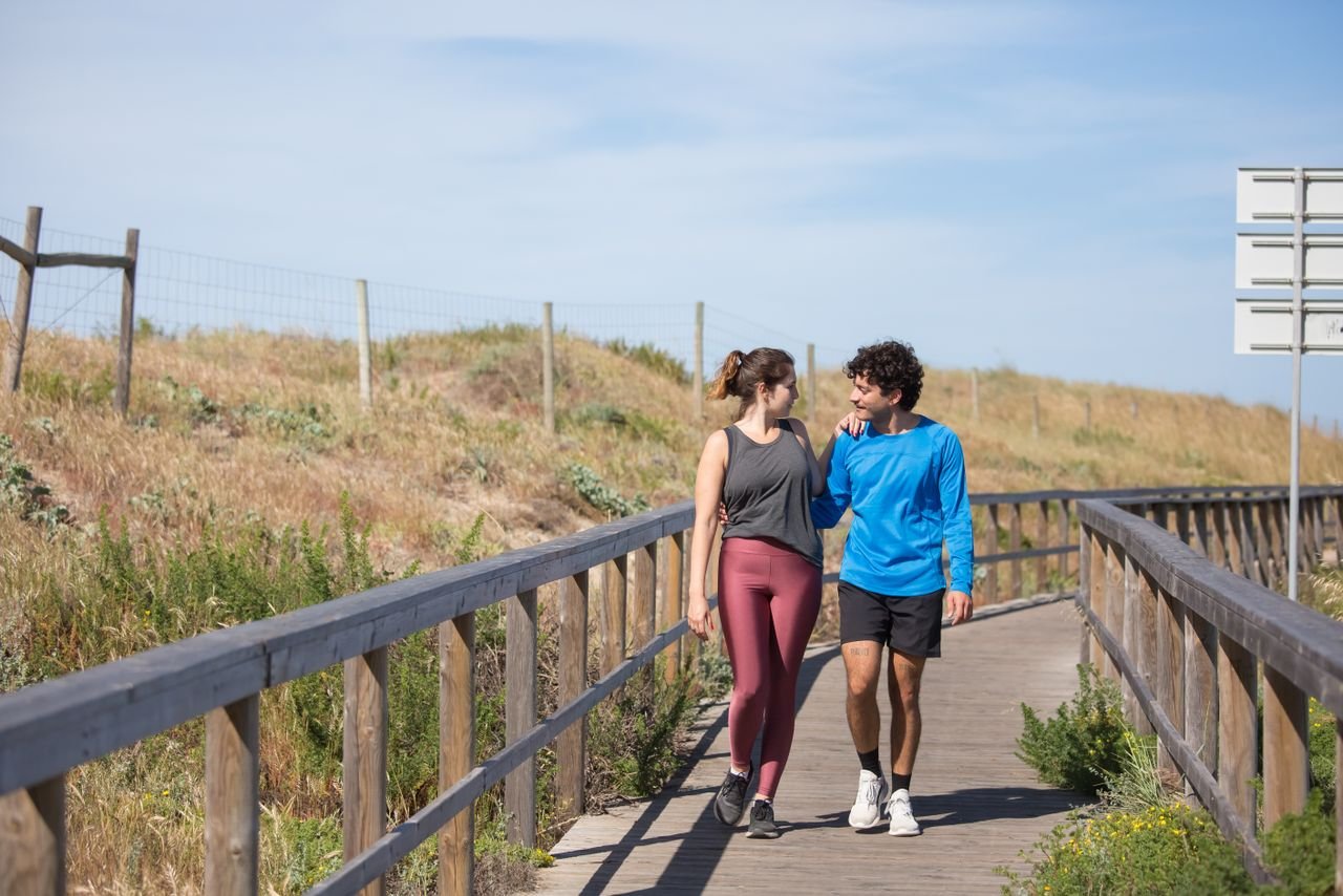A couple in activewear strolling together on a sunny boardwalk in a natural setting.